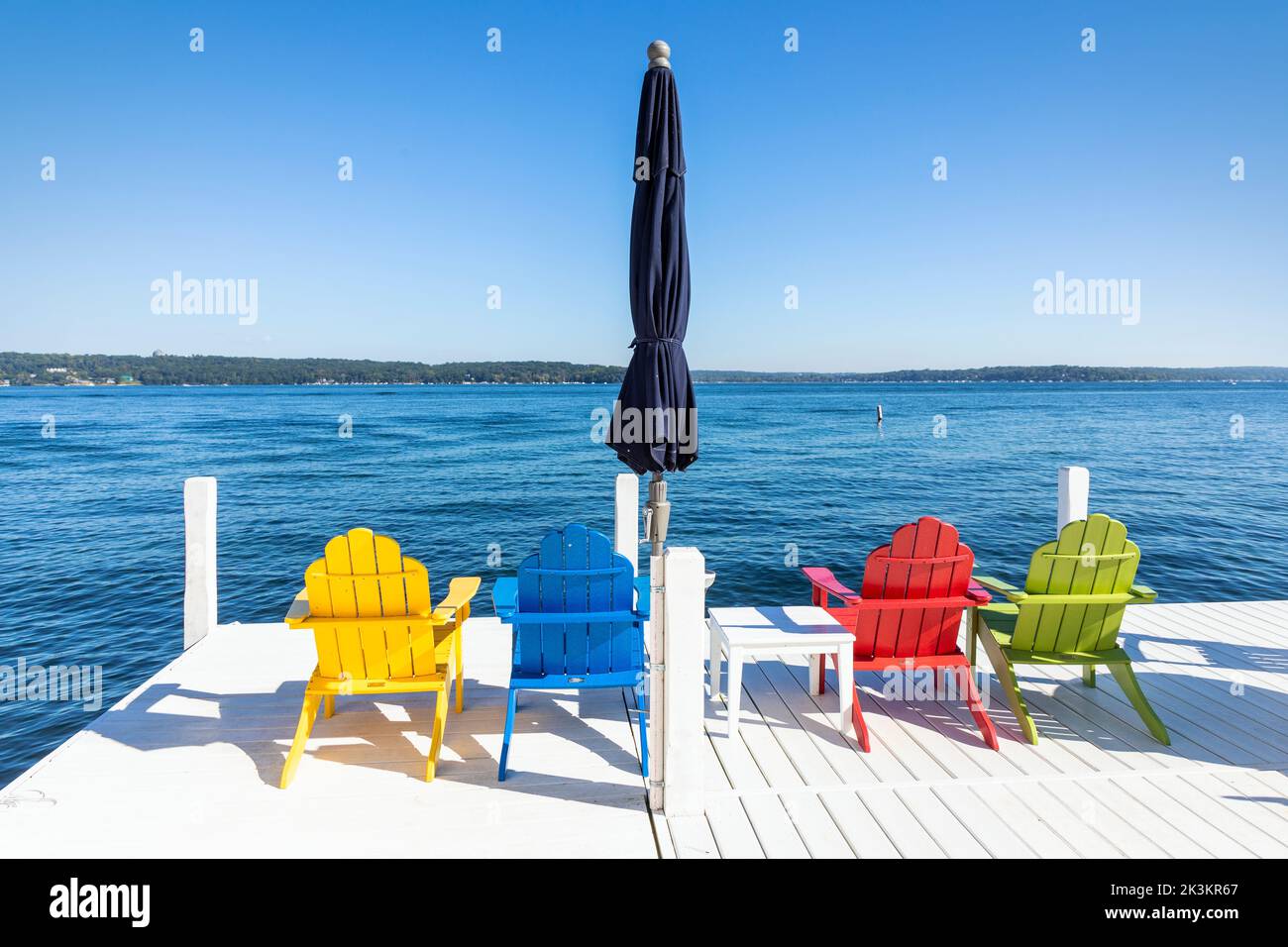 Vier farbige Holzstühle und ein blauer Sonnenschirm auf einem weißen Pier aus Holz, Genfersee in der Nähe von Fontana, Wisconsin, Amerika. Stockfoto