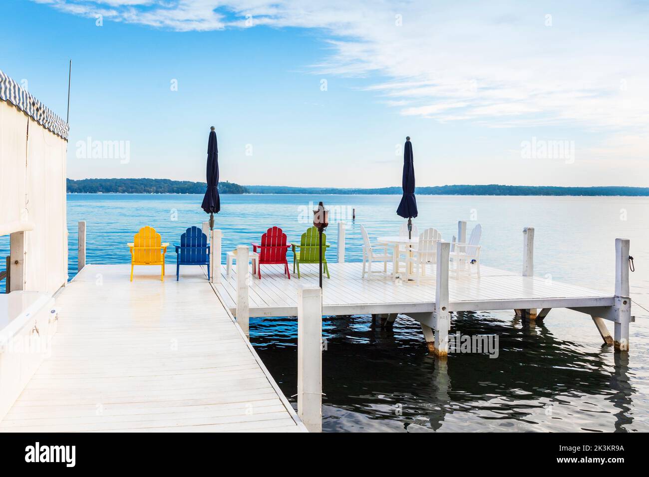 Vier farbige Holzstühle und ein blauer Sonnenschirm auf einem weißen Pier aus Holz, Genfersee in der Nähe von Fontana, Wisconsin, Amerika. Stockfoto