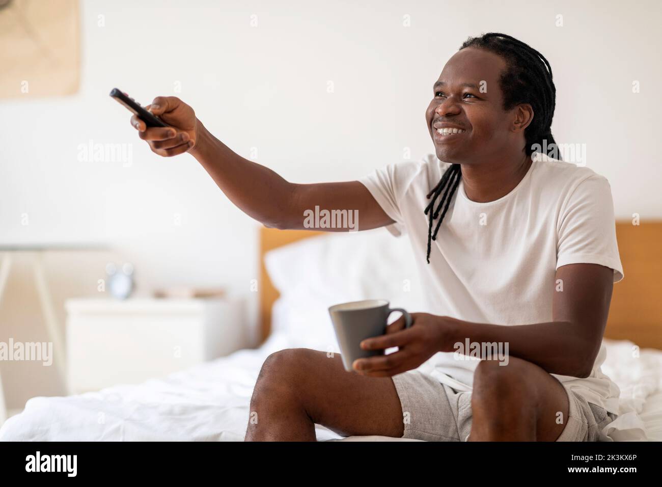 Lächelnder schwarzer Mann, der Kaffee trinkt und fernsieht, während er sich zu Hause entspannt Stockfoto