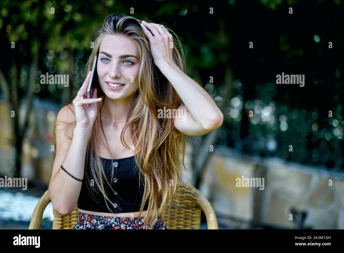 Natürliches Schönheitsporträt einer jungen Frau in den Zwanzigern mit langen Haaren und blauen Augen im Freien in einem Garten, der auf ihrem Mobiltelefon spricht. Lifestyle-Konzept. Stockfoto
