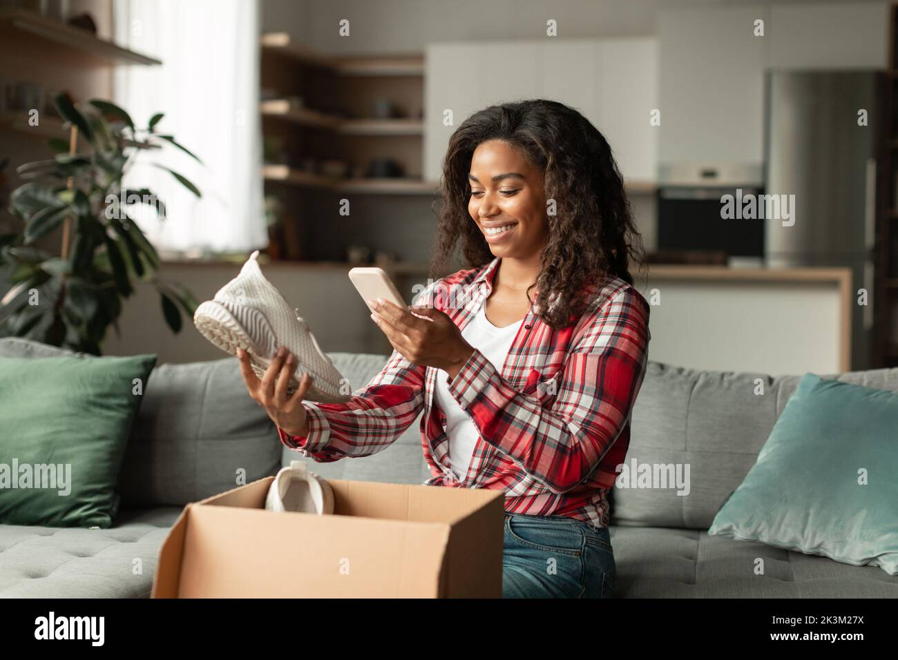 Die junge afroamerikanische Dame öffnet die Box, fotografiert Schuhe auf dem Smartphone, genießt Online-Shopping Stockfoto