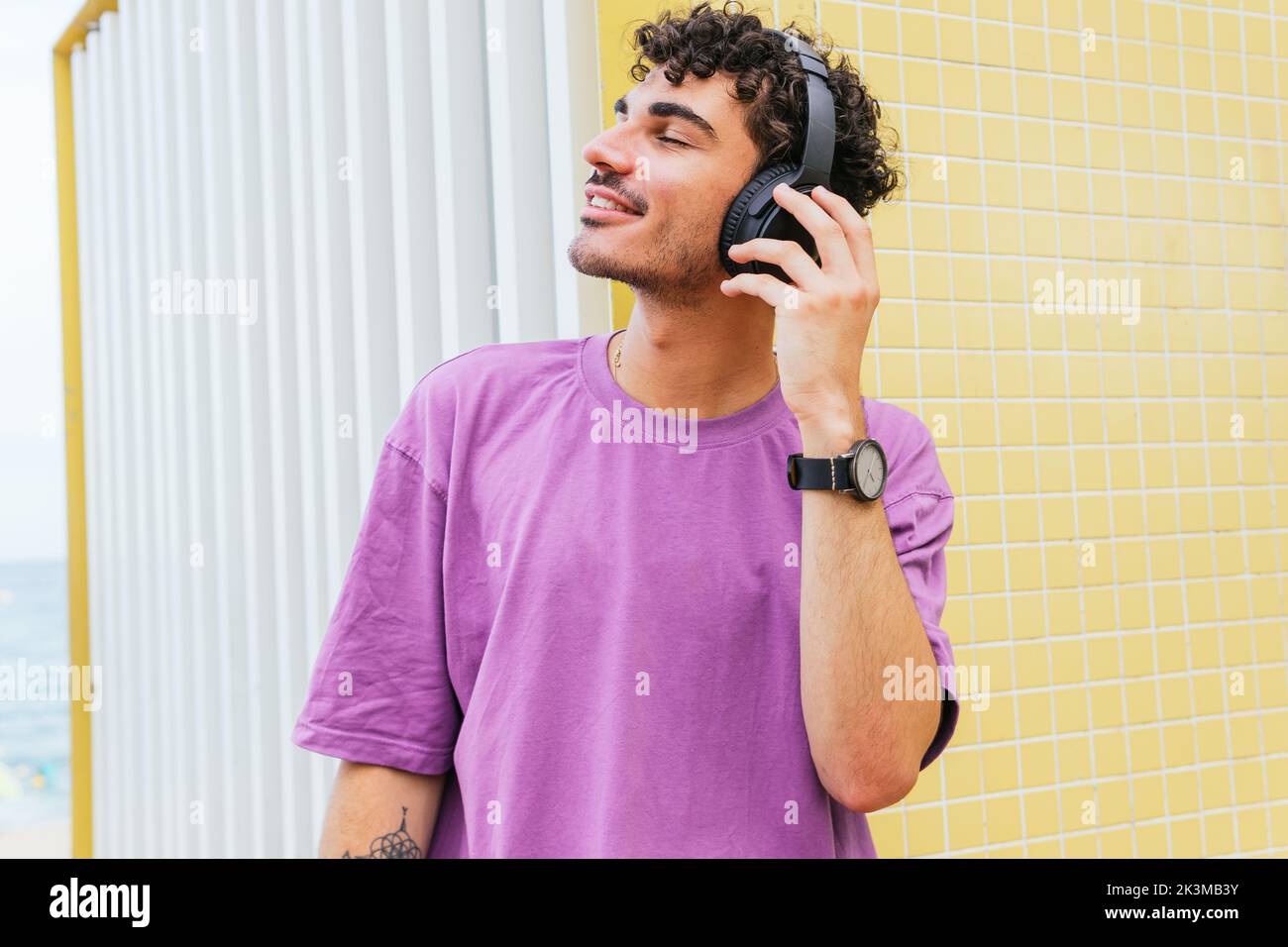 Fröhlicher hispanischer Mann im violetten T-Shirt, der kabellose Kopfhörer anpasst und mit geschlossenen Augen lächelt, während er Lieblingsmusik in der Nähe von gelb gefliestem Wal hört Stockfoto