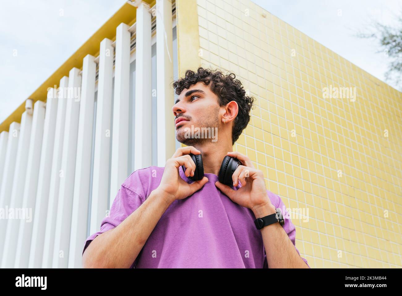 Hispanic-Mann in lila T-Shirt Anpassung drahtlose Kopfhörer wegschauen, während Sie Lieblingsmusik in der Nähe gelb gefliesten Wand am Wochenende am b hören Stockfoto