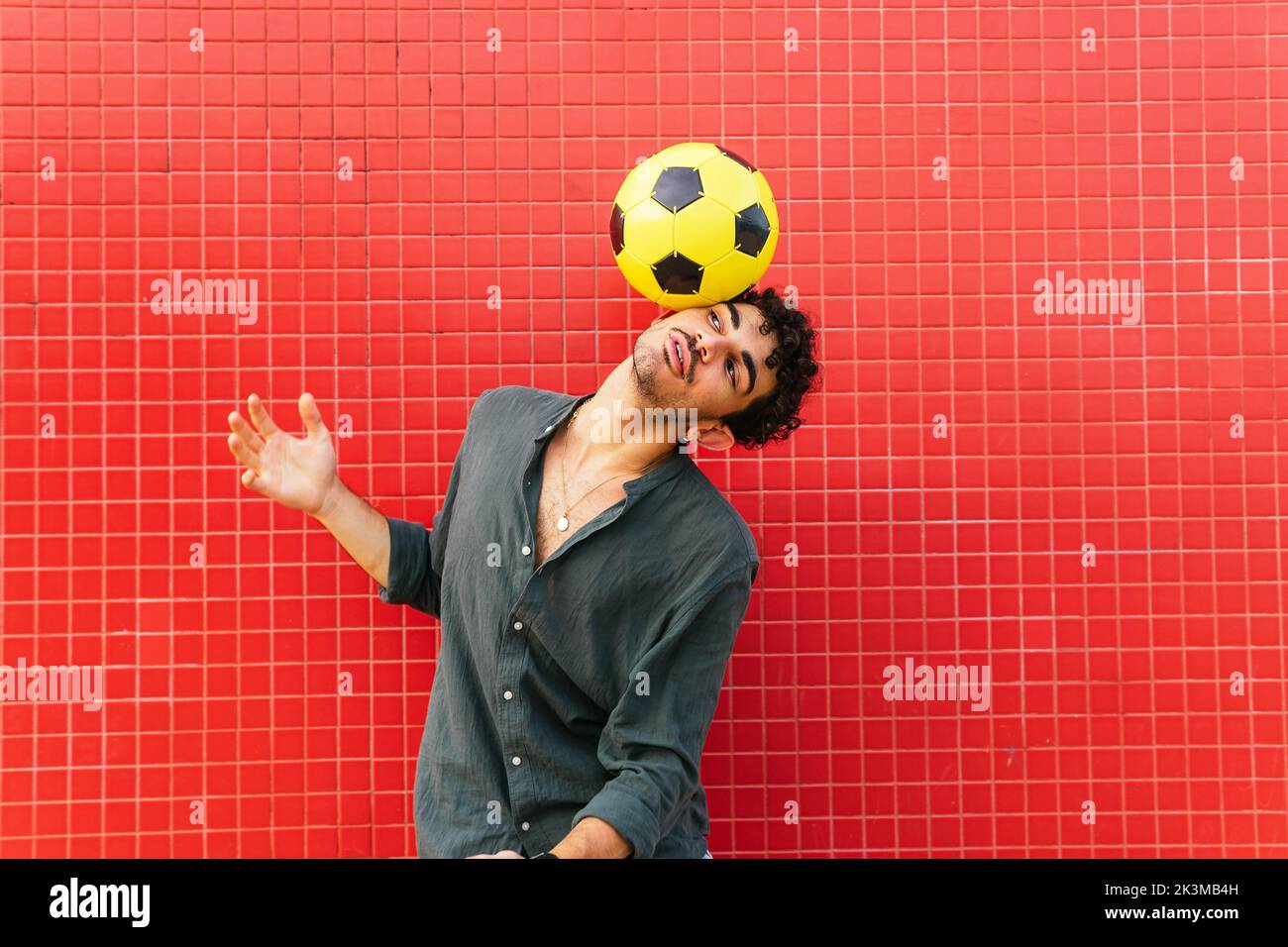 Junger hispanischer Mann im schwarzen Hemd balanciert gelben Fußballball am Kopf gegen geflieste rote Wand auf der Straße Stockfoto