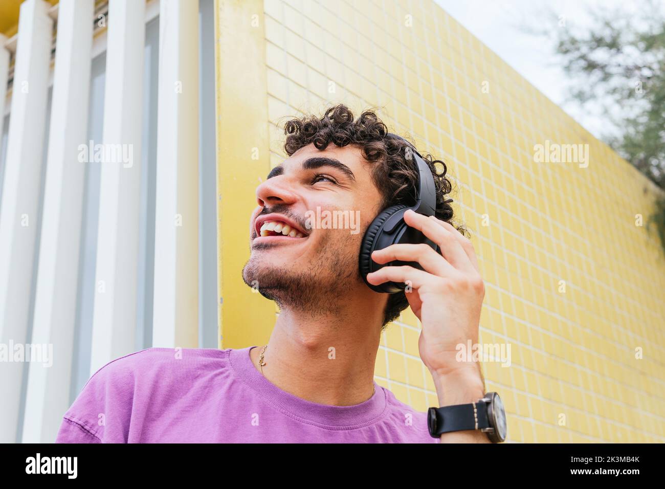 Fröhlicher Hispanic-Mann im lila T-Shirt, der kabellose Kopfhörer anpasst und lächelnd wegschaut, während er an der gelb gefliesten Wand Lieblingsmusik hört Stockfoto