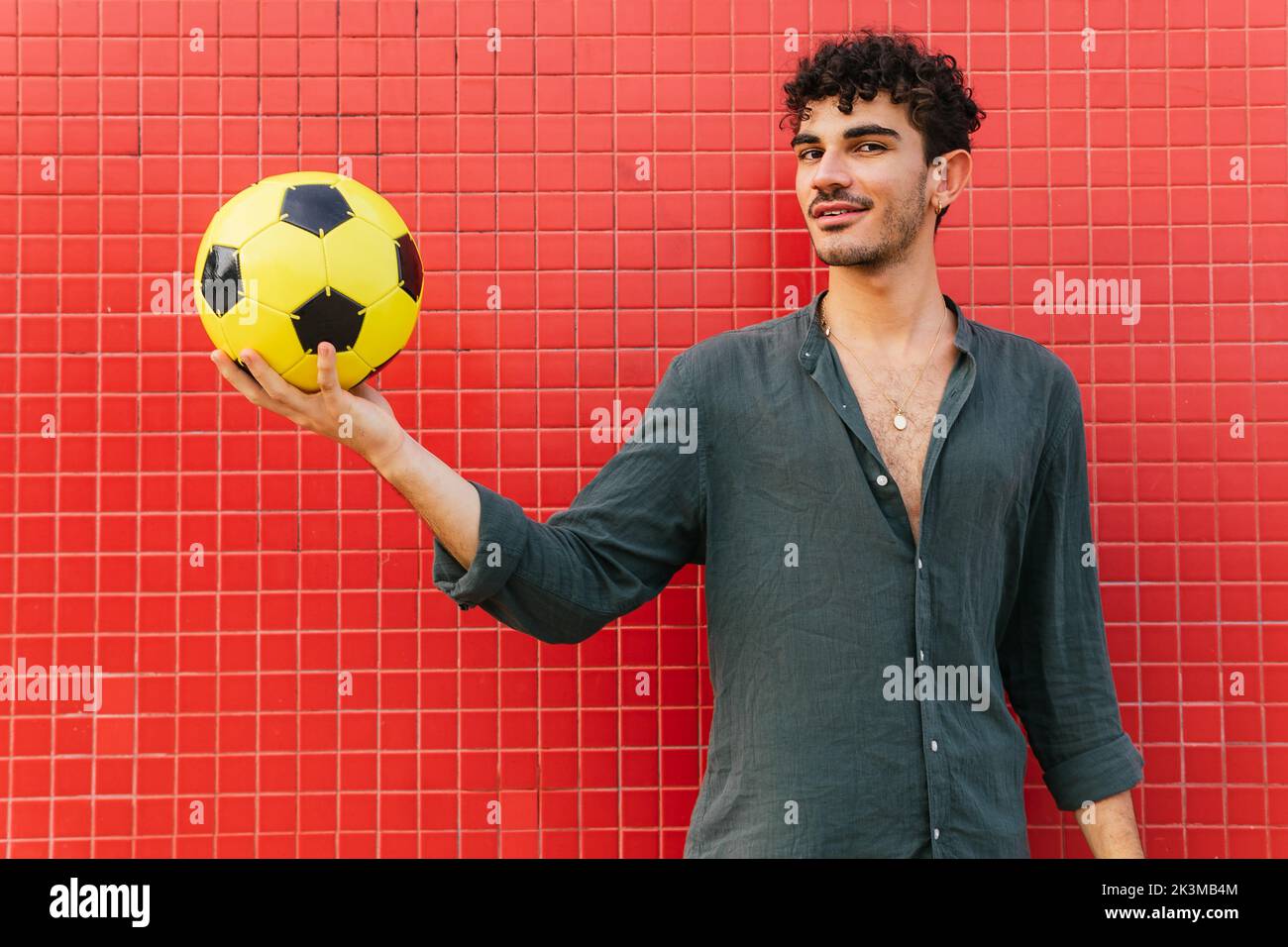 Junger Hispanic-Mann in schwarzem Hemd, der den gelben Fußballball auf der Hand hält und auf der Straße gegen die geflieste rote Wand steht Stockfoto