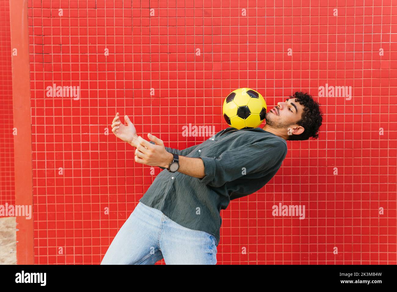 Seitenansicht eines jungen hispanischen Mannes in schwarzem Hemd, der auf der Brust den gelben Fußballball gegen die geflieste rote Wand auf der Straße balanciert Stockfoto