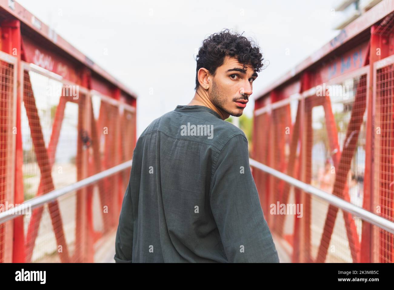 Rückansicht eines jungen hispanischen Mannes in schwarzem Hemd, der auf einer Brücke geht und die Kamera über die Schulter betrachtet, während er auf einer Fußgängerbrücke am Strand läuft Stockfoto