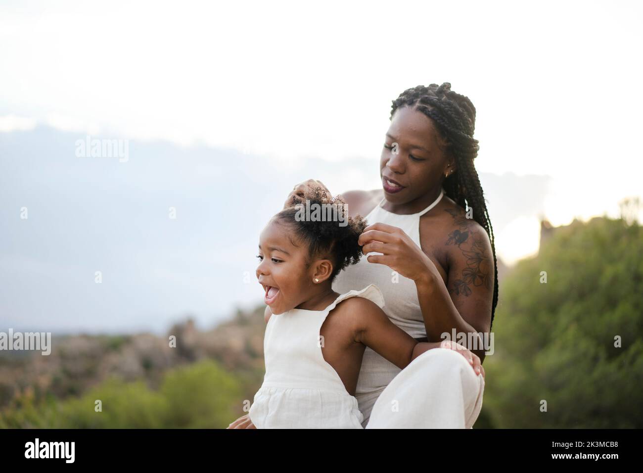 Schwarze Mutter mit Afro-Zöpfen, die die Haarbrötchen der glücklichen Tochter anpassen, während sie zusammen das Sommerwochenende in der Natur verbringt Stockfoto