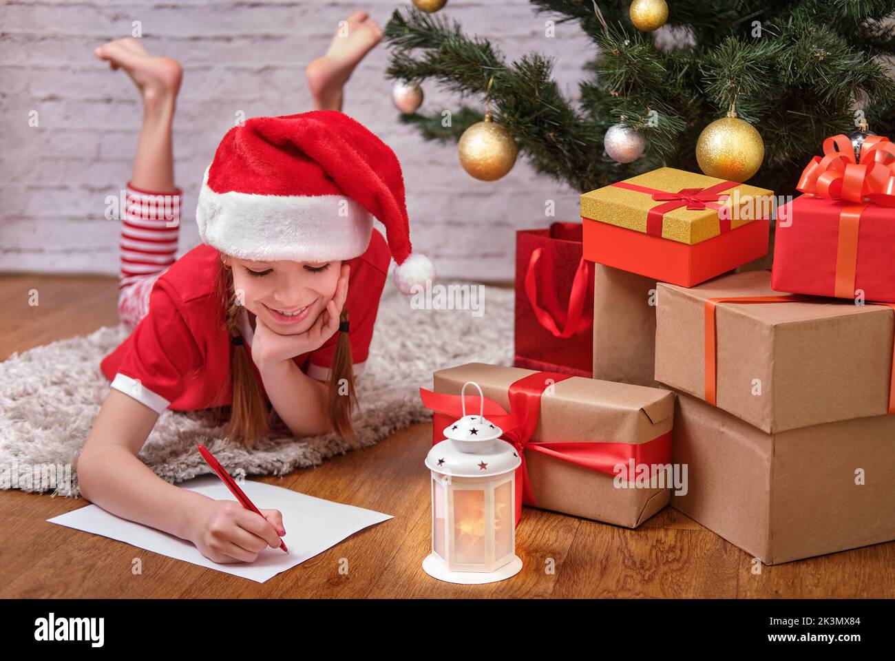 Ein fröhlich lächelndes Weihnachtsmädchen in einem Weihnachtshut schreibt einen Brief an den Weihnachtsmann, der auf dem Boden in der Nähe des Weihnachtsbaums und der Weihnachtsgeschenke liegt. Stockfoto