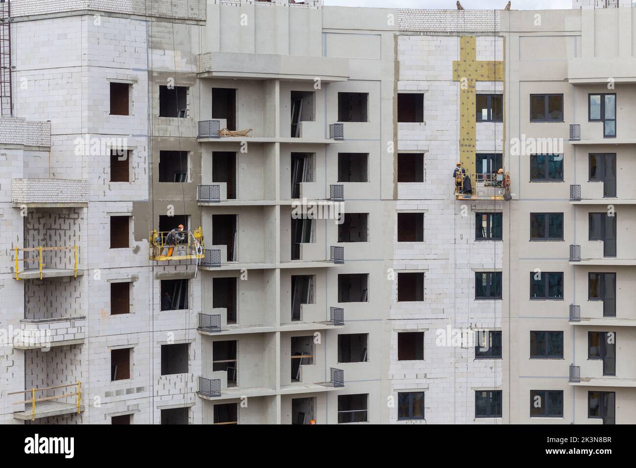 Der Prozess der Isolierung von Außenwänden in einem mehrstöckigen Gebäude im Bau. Die Arbeiter in einer orangen Konstruktionswiege befestigen die Mineralwolle s Stockfoto