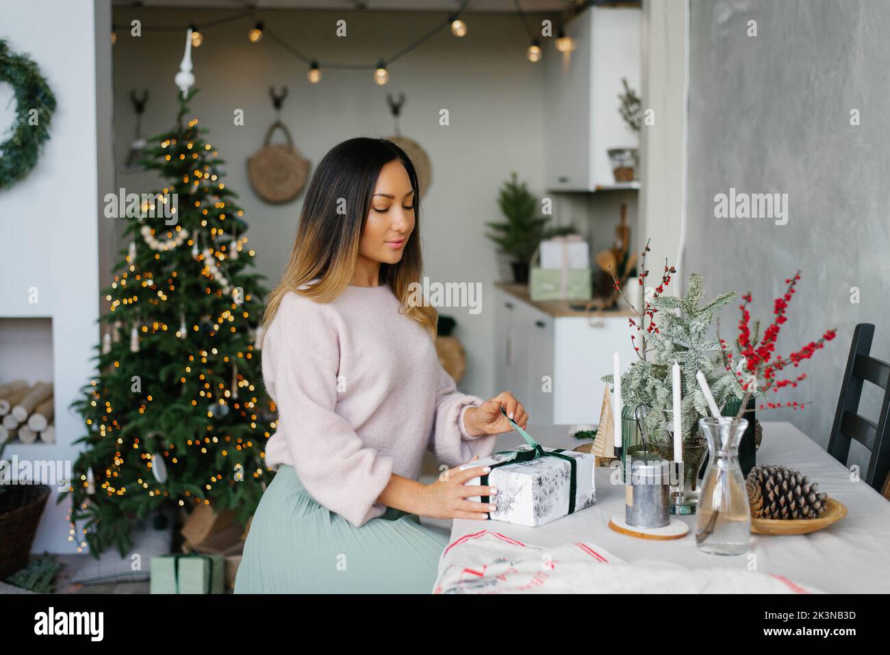 Eine stilvolle Frau, die an einem festlichen Tisch sitzt, öffnet ein Weihnachtsgeschenk Stockfoto