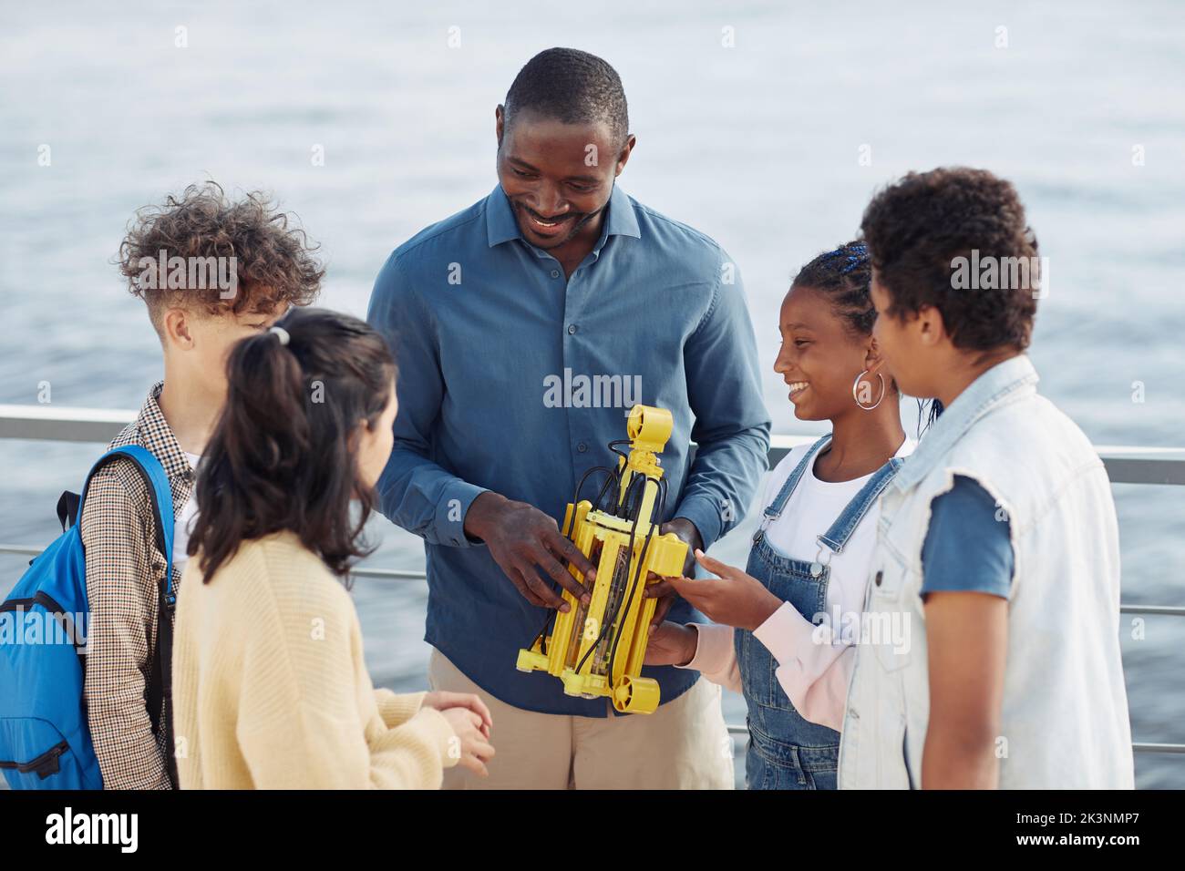 Porträt eines lächelnden männlichen Lehrers, der einer Gruppe von Kindern, die während des Ingenieursvorstehens im Freien im Kreis stehen, ein Robotermodell zeigt Stockfoto