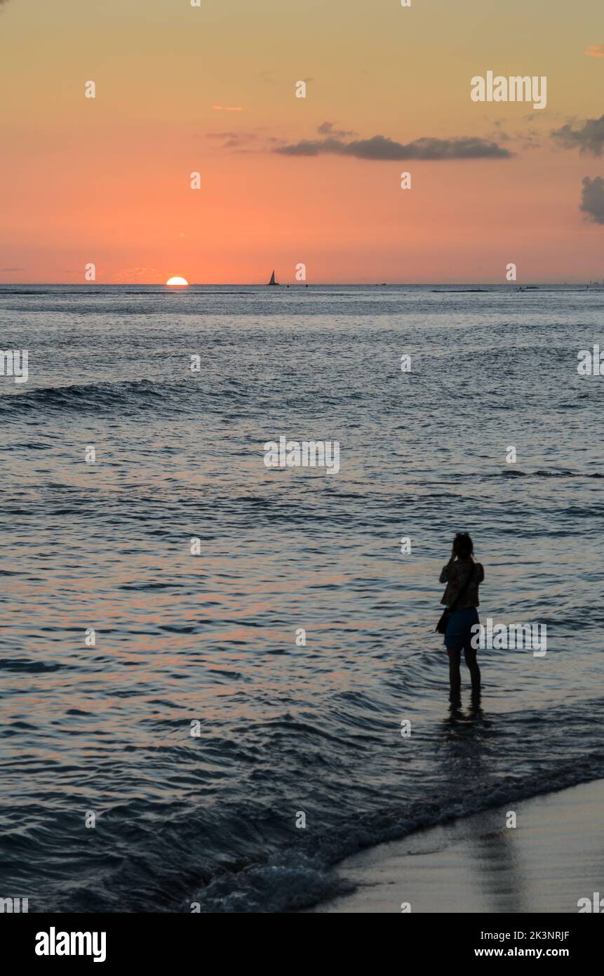 Eine alleinreisende Frau am Waikiki Strand in Hawaii, die ein Foto vom Sonnenuntergang über dem Pazifischen Ozean gemacht hat Stockfoto