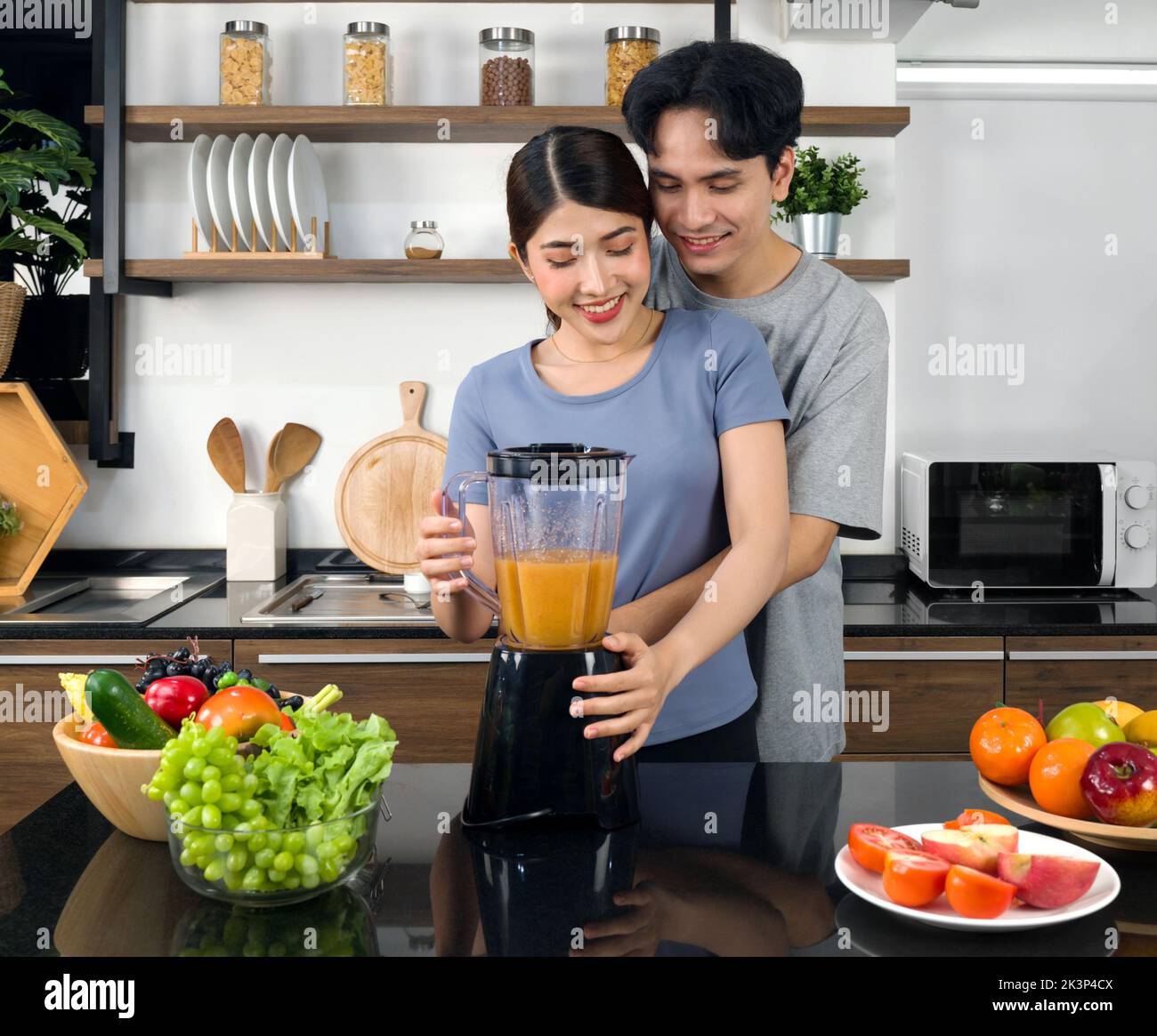Junge asiatische Frau mit Mischmaschine mischen Obst und Gemüse in Smoothie. Ihr Freund kuschelt von hinten. Die Küchentheke voller Vari Stockfoto