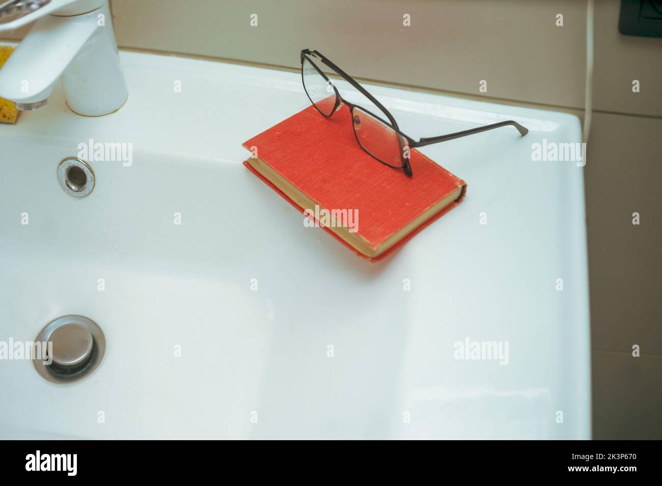 Ein Buch und eine Brille auf einem Waschbecken im Badezimmer, Nahaufnahme im Innenbereich, flacher innenfrauenabfluss Stockfoto
