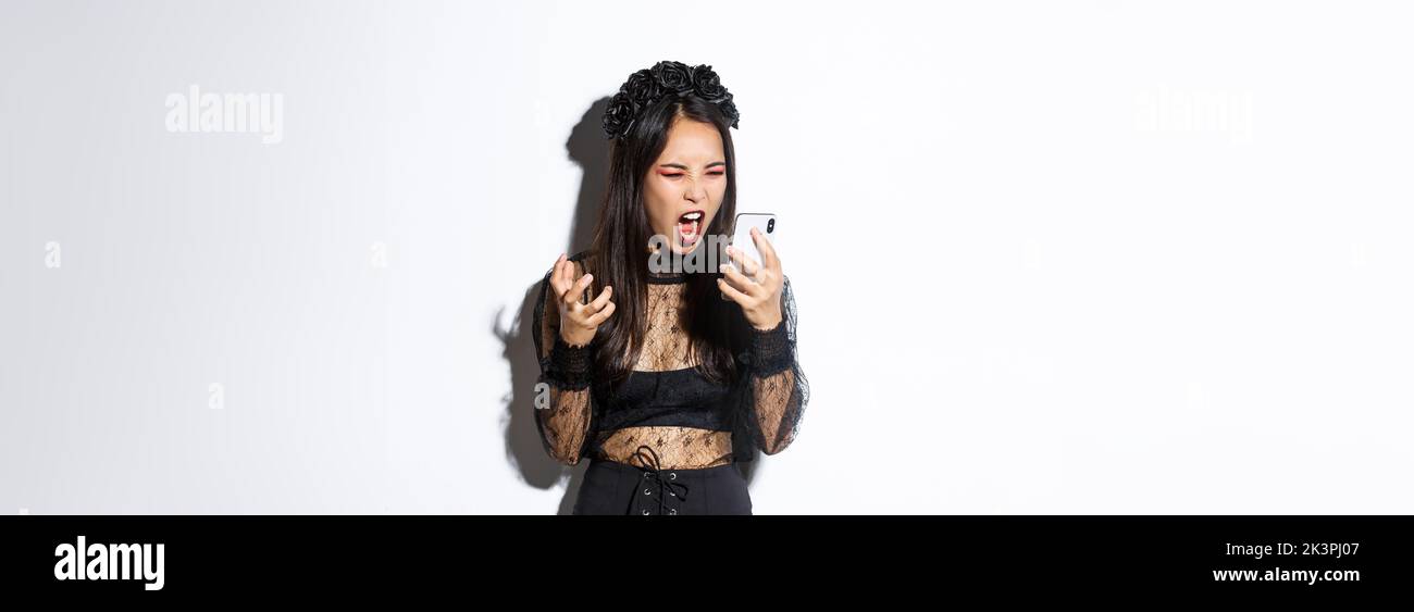 Porträt der wütenden asiatischen Frau in halloween Kostüm verrückt, schreien auf Handy und Grimacing wütend, stehend über weißem Hintergrund Stockfoto