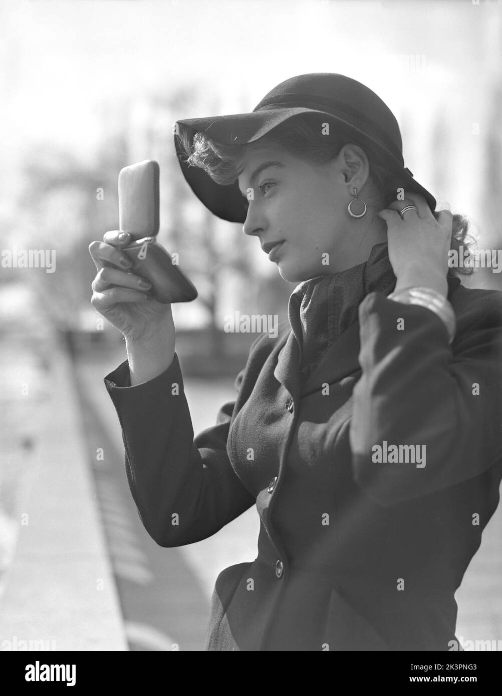 Make-up in der 1950s. Eine junge Frau hält einen Taschenspiegel und fixiert ein Detail in ihren Haaren. Das Model trägt eine Jacke mit einer sichtbaren dünnen Taille, einem passenden Hut und einem Schal um den Hals. Schweden 1952 Kristoffersson Ref 38K-7 Stockfoto