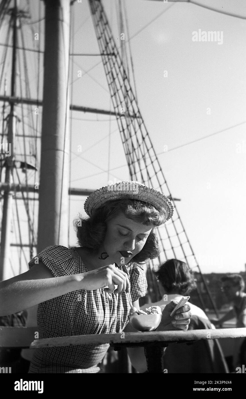 In der 1940s. Eine junge Frau besucht die Hauptstadt Schwedens, Stockholm, und genießt die Sonne und eine Pause bei der Besichtigung der Stadt. Sie trägt Sommerkleid und Hut und isst Eiscreme auf dem Segelschiff AF Chapman, das vor Anker liegt und als Café und Hostel fungiert. Stockholm Schweden 1949. Kristoffersson Ref. AO30-4 Stockfoto