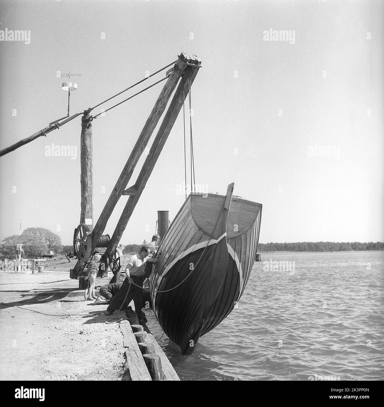 1950s Boot. Was wie ein dramatisches Ereignis aussieht, ist in Wirklichkeit ein kontrollierter Start eines hölzernen Segelbootes. Der alt aussehende Kran lässt das Boot im Wasser schön und einfach runter. Solvesborg Schweden 1954 Kristoffersson Ref. BX28-6 Stockfoto