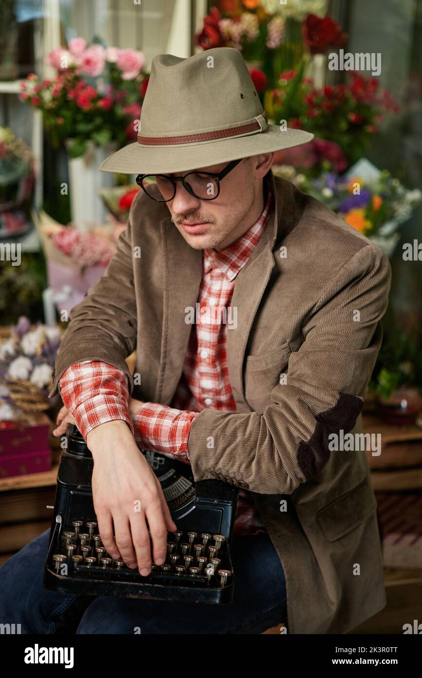 Attraktive kaukasische männliche Person in Brillen und Hut sitzend mit alter Schreibmaschine im Blumengeschäft denken. Dichter oder Schriftsteller Arbeitskonzept. Hochwertiges vertikales Bild Stockfoto