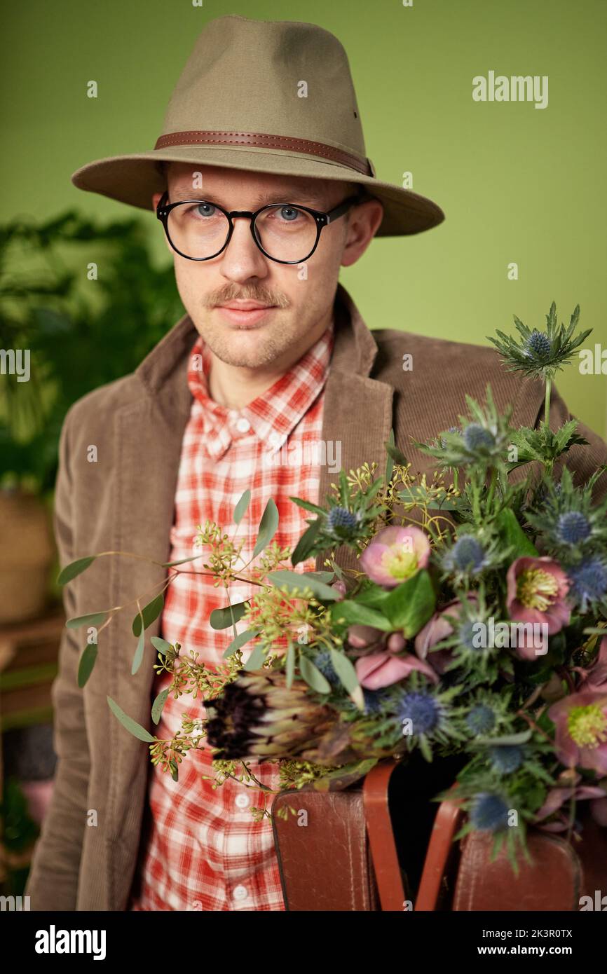 Niedliche altmodische männliche Person in Brillen, Samtjacke und Hut mit Blumenstrauß und Aktentasche im Blumenladen bleiben. KameraHochwertiges vertikales Bild Stockfoto