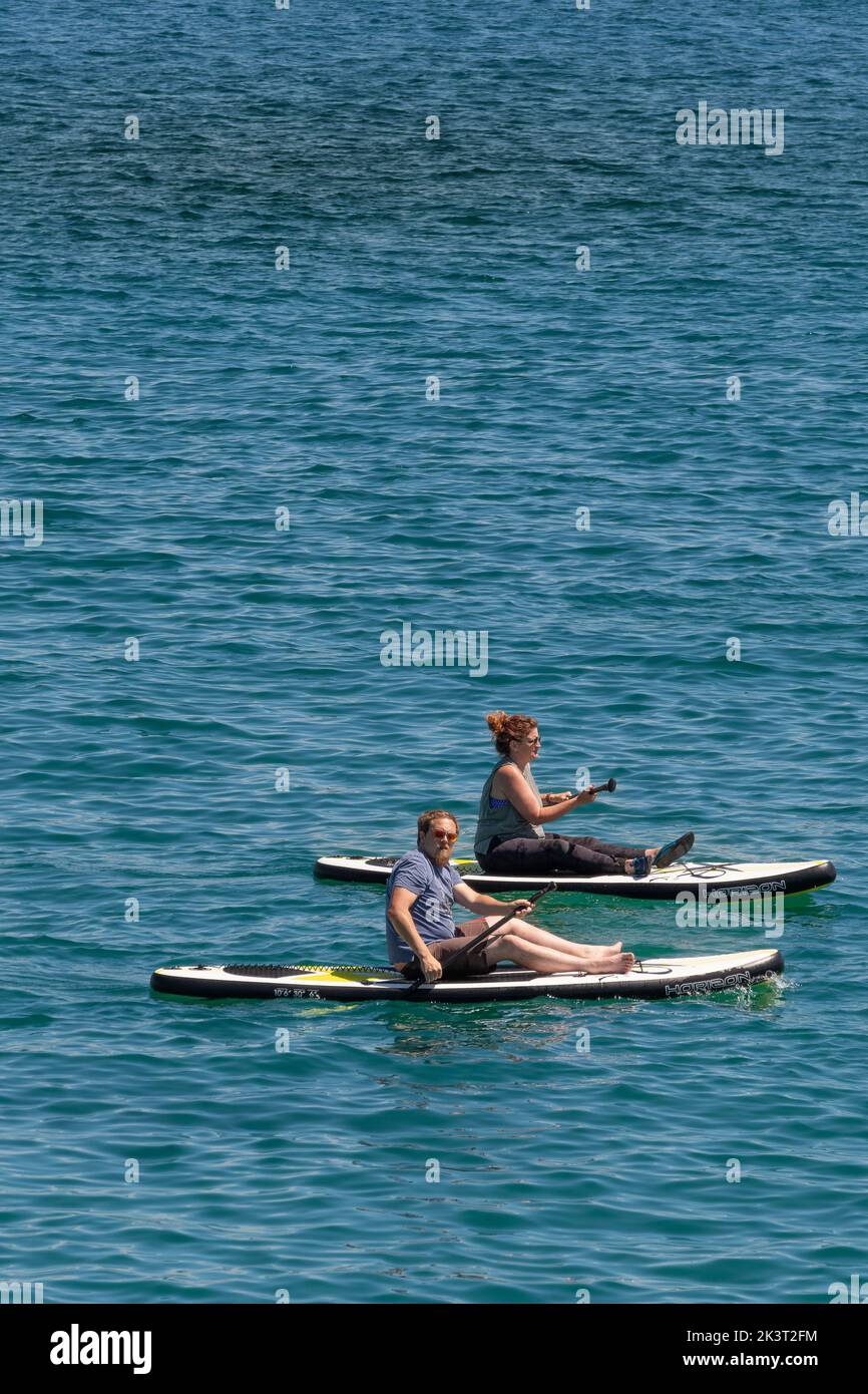 Zwei Urlauber sitzen auf Stand Up Paddleboards in Newquay Bay in Cornwall in England im Vereinigten Königreich. Stockfoto