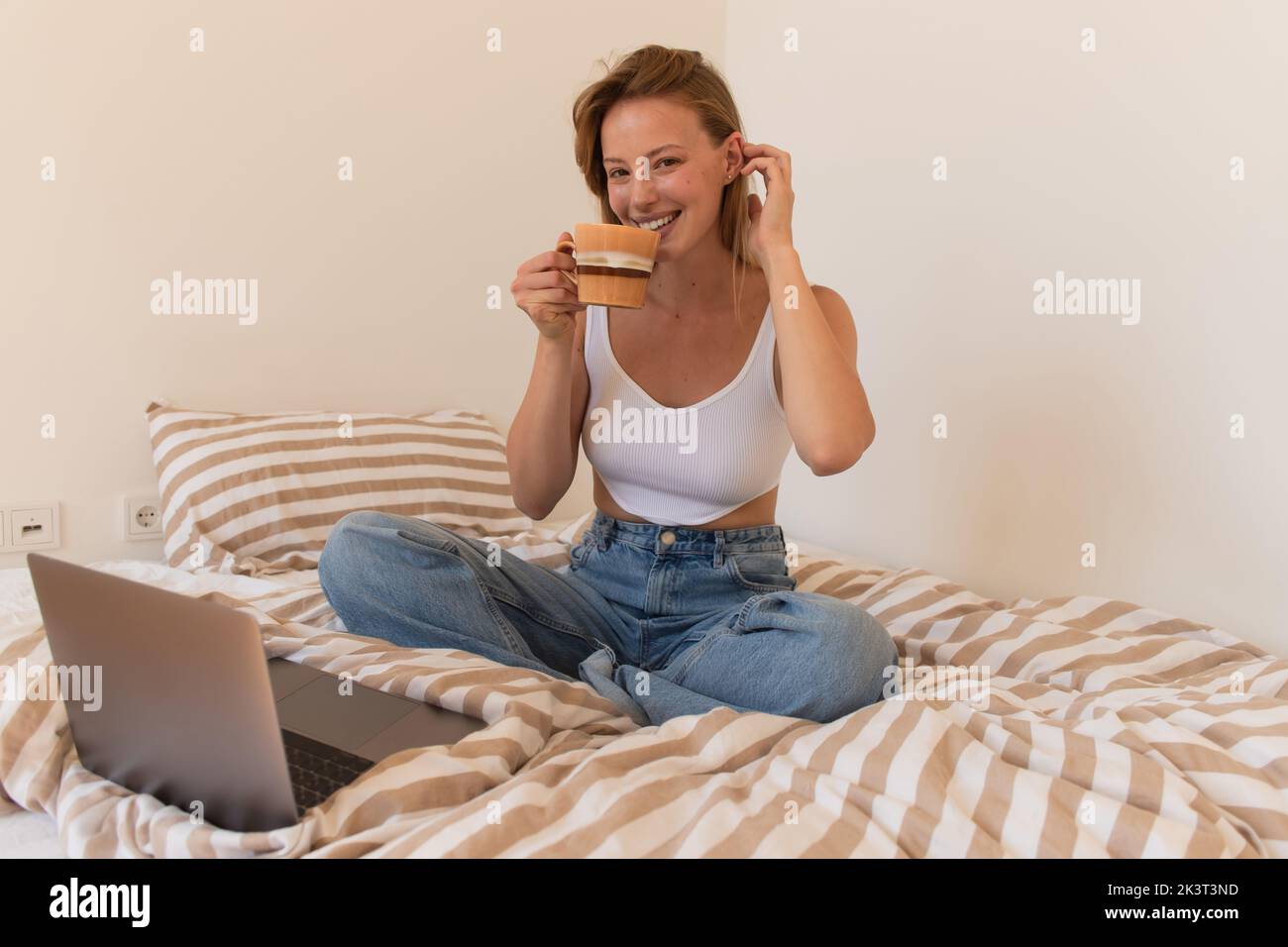 Junger Freiberufler lächelt der Kamera zu, während er Kaffee in der Nähe des Laptops auf dem Bett trinkt, Stockbild Stockfoto
