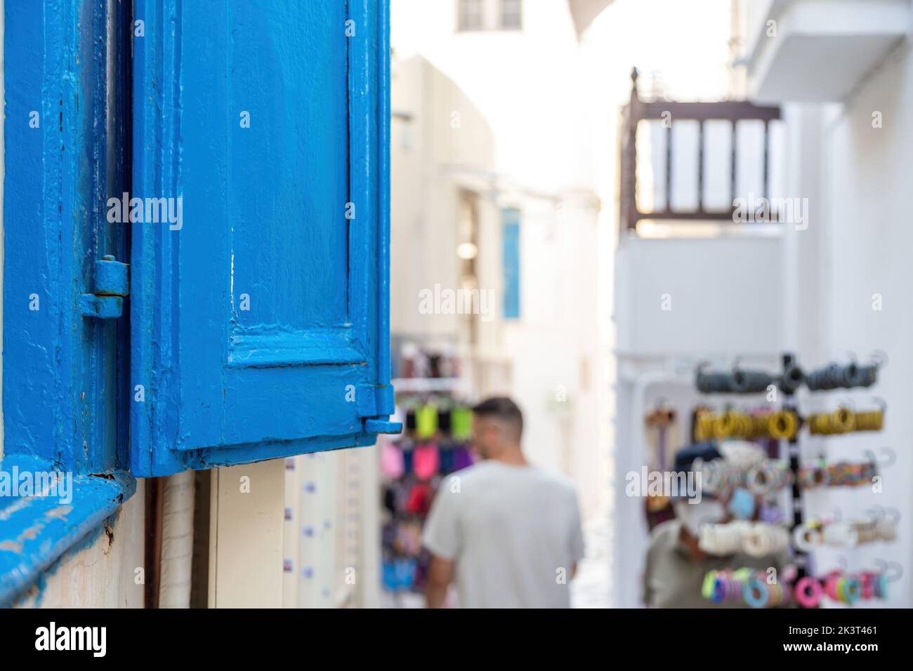 Insel Naxos, Kykladen Griechenland. Blick auf einen blauen, offenen Holzverschluss und einen verschwommenen Mann, der sich ein Souvenir im Freien zwischen traditionellen weiß getünchten Gebäuden anschaut. Stockfoto
