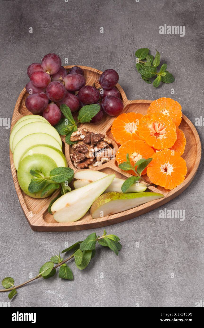 Herzförmiger Holzteller mit Früchten und Nussbaum auf grauem Hintergrund Stockfoto