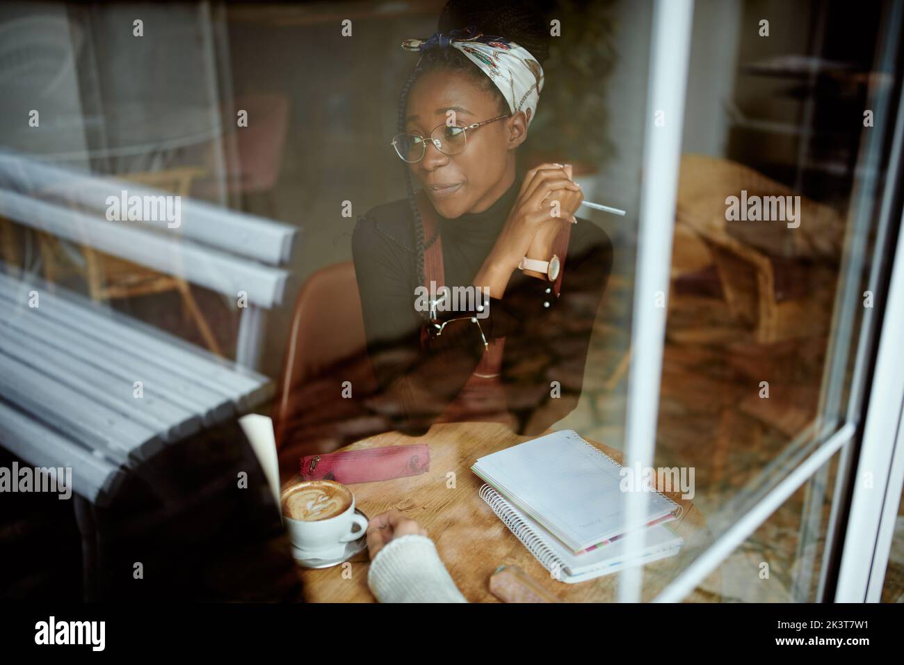 Ein schickes afrikanisches College-Mädchen sitzt in der Cafeteria und studiert für eine Prüfung. Das Bild wurde von außen aufgenommen. Ein Mädchen im Studentenaustausch sitzt Stockfoto