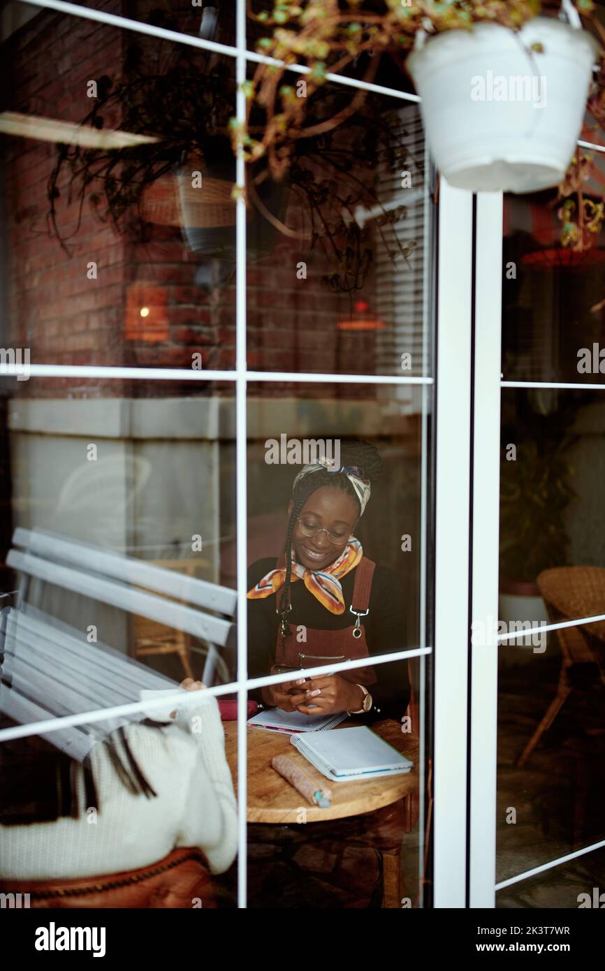 Blick durch das Fenster auf die junge afroamerikanische Hipster-Frau, die im Internetcafe studiert. Stockfoto