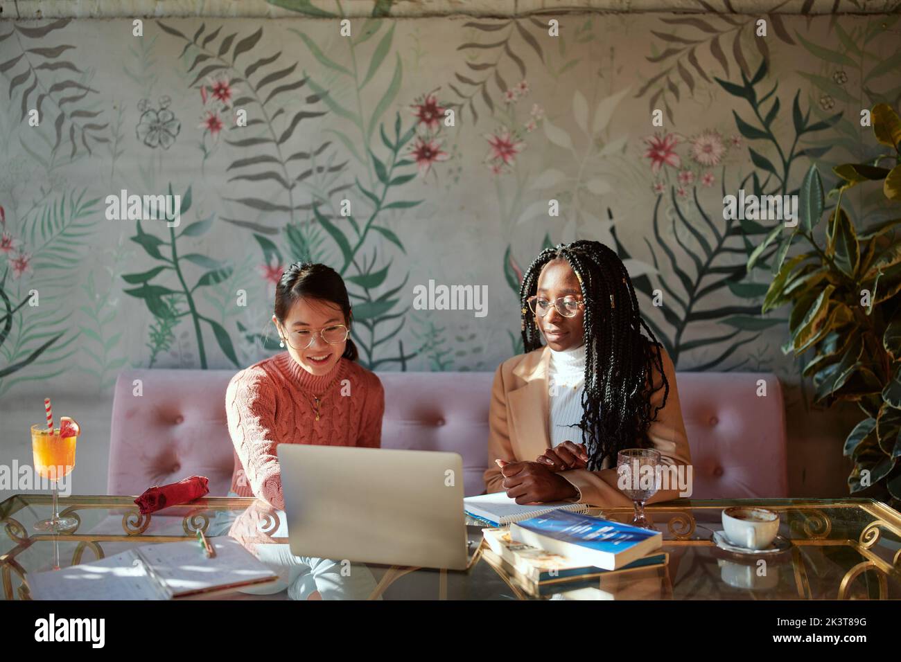 Zwei intelligente College-Mädchen sitzen in einem Café mit Büchern auf einem Tisch und nutzen einen Laptop für einen Online-Kurs. Mädchen, die in einem Coffee Shop studieren. Stockfoto