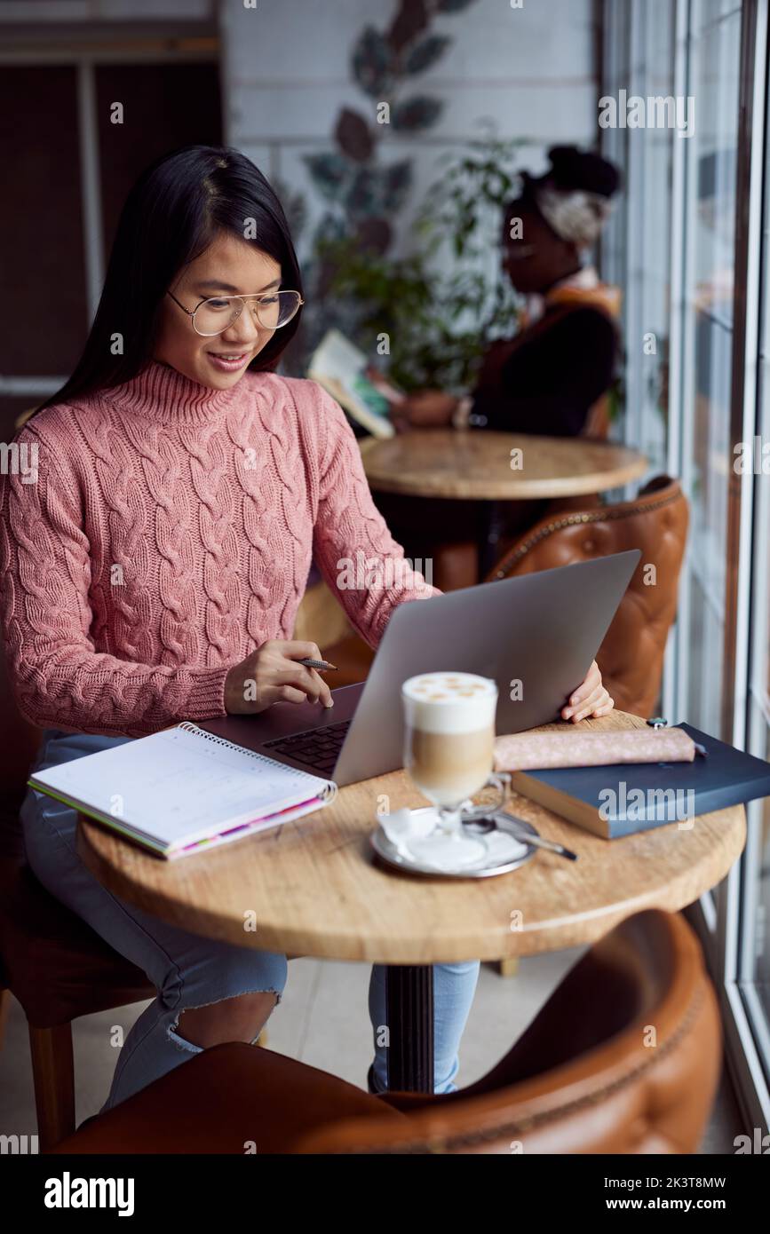 Ein glückliches multikulturelles College-Mädchen sitzt in einem Café und macht Aufgaben auf einem Laptop. Stockfoto