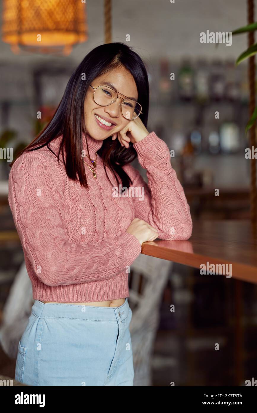 Eine asiatische junge Frau, die sich im Café auf den Tisch lehnt und die Kamera anlächelt. Stockfoto