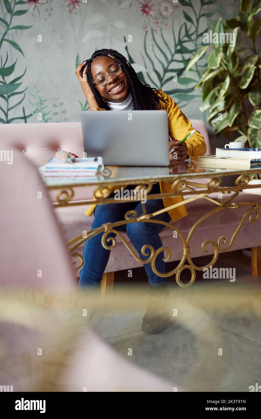 Ein lächelndes afrikanisches College-Mädchen sitzt in einem Café und beendet das Schulprojekt auf ihrem Laptop. Stockfoto