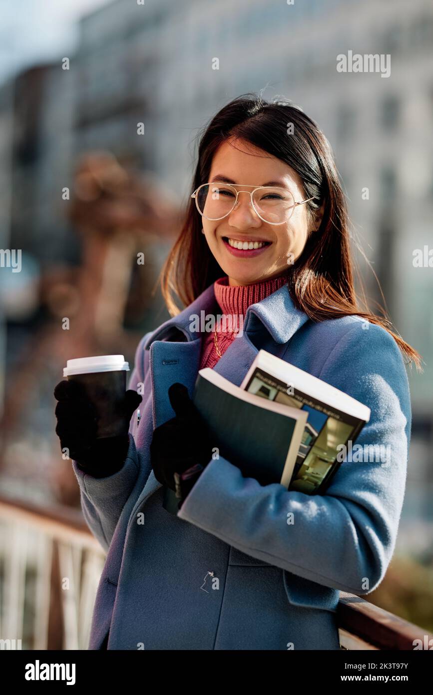 Portrait eines glücklichen asiatischen Mädchens hält Bücher und Kaffee zum Mitnehmen draußen bei kaltem Wetter. Stockfoto