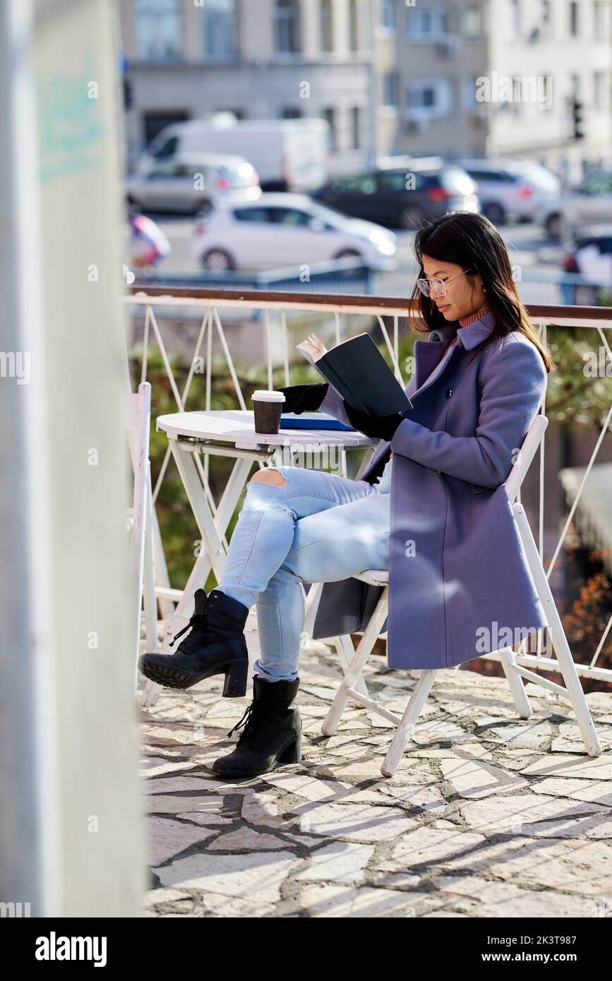 Ein asiatisches Mädchen sitzt bei kaltem Wetter auf dem Balkon einer Cafeteria und liest ein Buch. Stockfoto