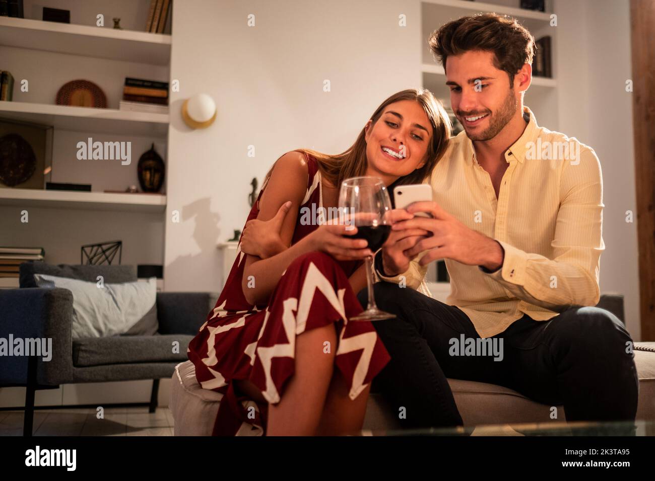 Ein fröhlicher junger Mann teilt Fotos auf dem Handy mit einer Frau, die sich auf seiner Schulter lehnt und zu Hause auf dem Sofa sitzt Stockfoto Ein fröhlicher junger Mann teilt Fotos auf dem Handy mit einer Frau, die sich auf seiner Schulter lehnt und zu Hause auf dem Sofa sitzt Stockfoto