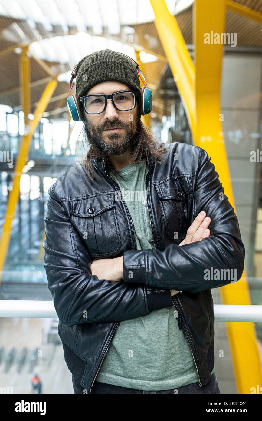 Hübscher bärtiger Hipster mit Brille und Kopfhörer in schwarzer Lederjacke und Strickmütze, der im Flughafen hal auf die Kamera schaut Stockfoto