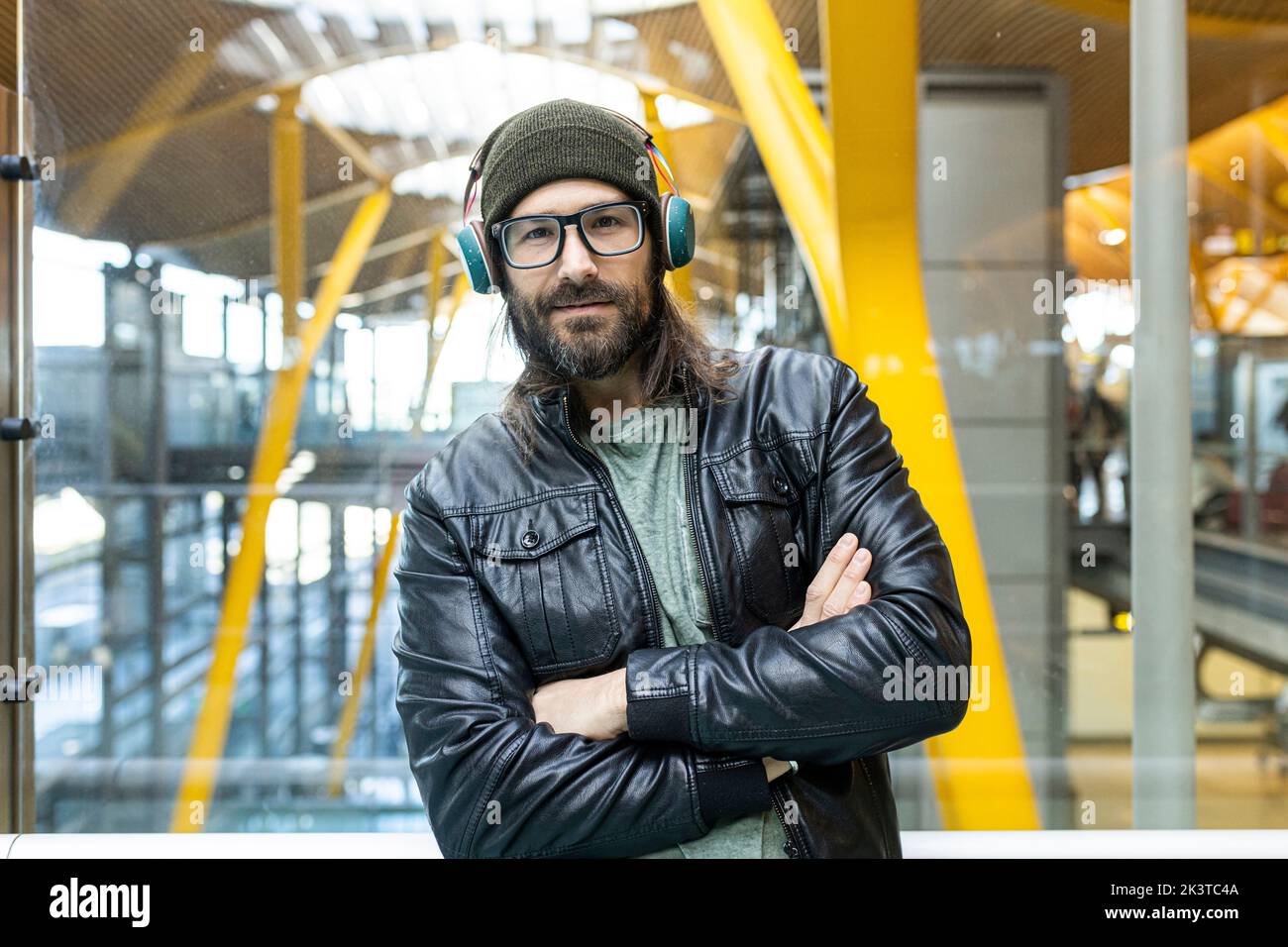 Hübscher bärtiger Hipster mit Brille und Kopfhörer in schwarzer Lederjacke und Strickmütze, der im Flughafen hal auf die Kamera schaut Stockfoto