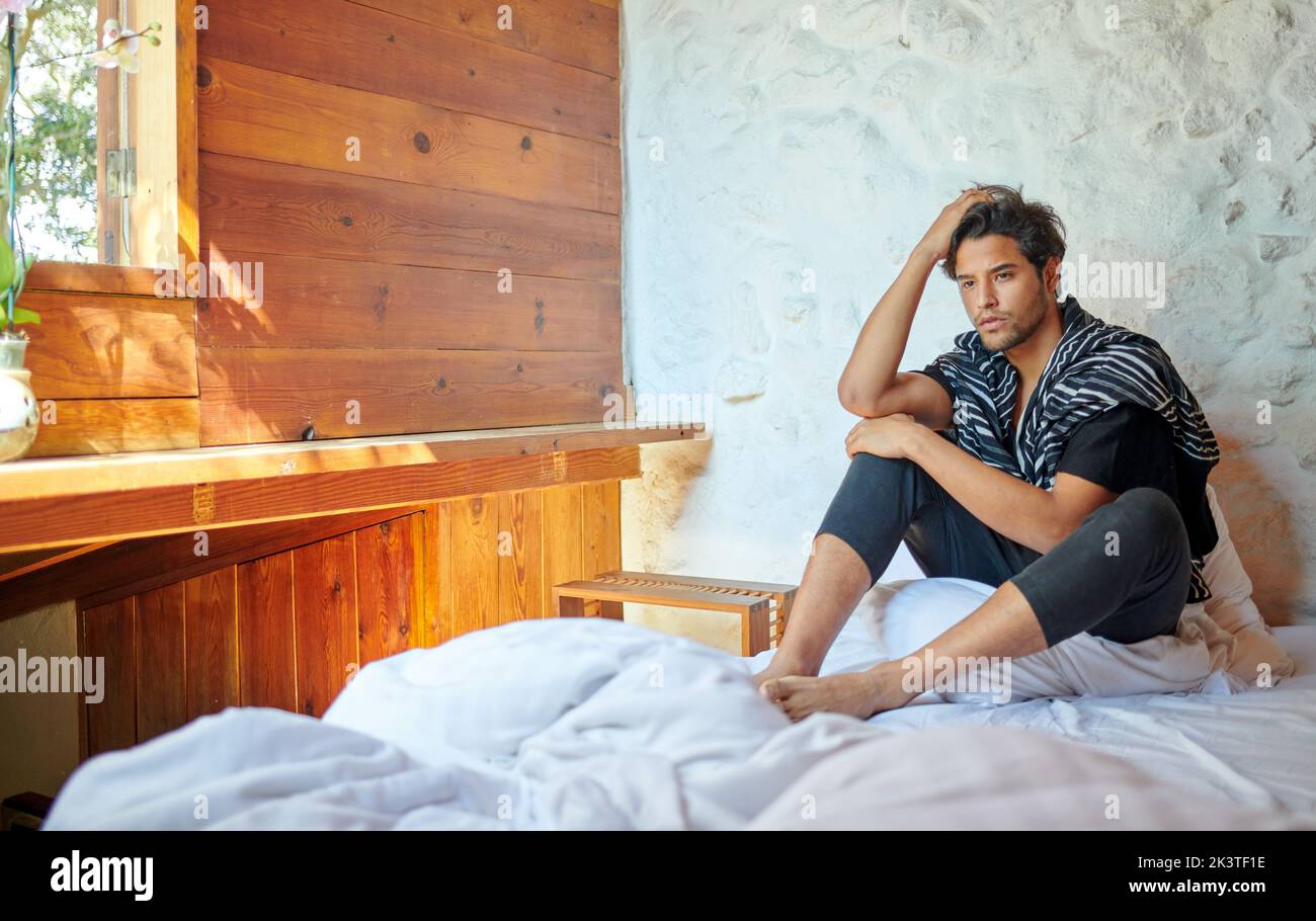 Junger nachdenklicher hispanischer Mann in stilvoller Kleidung, der den Kopf berührt, während er sich zu Hause auf dem geknickten Bettlaken nach vorne freut Stockfoto