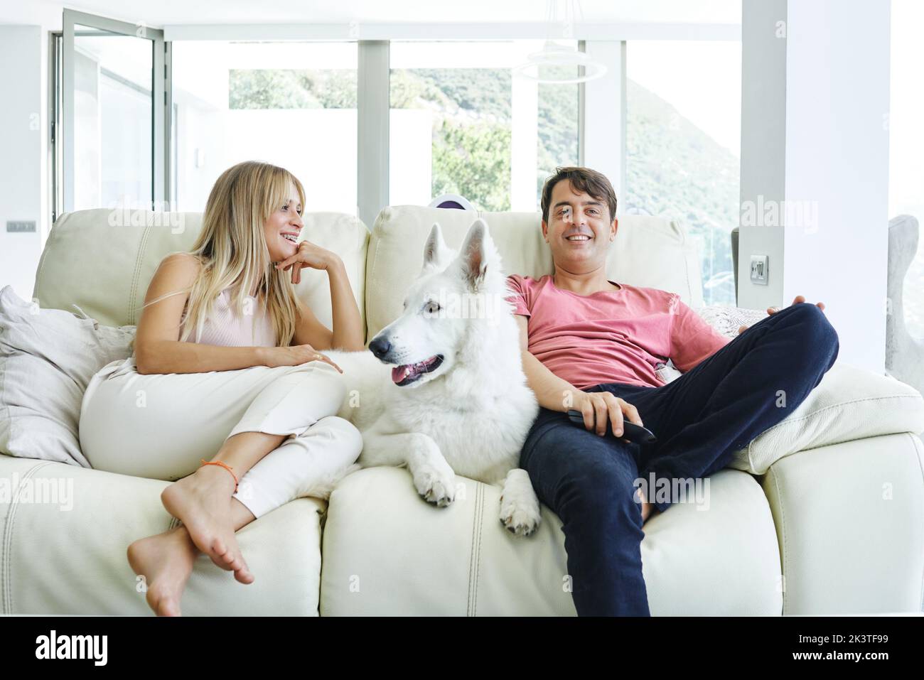 Paar ruhen sich mit dem weißen Schweizer Schäferhund auf der Couch aus, während sie das Wochenende gemeinsam genießen und zu Hause Fernsehen Stockfoto