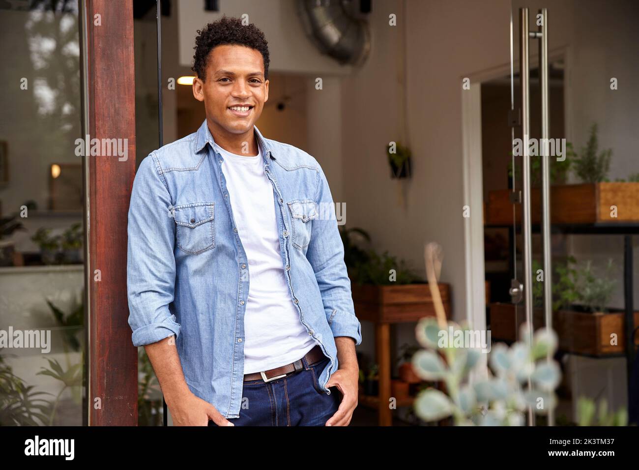 Lateinamerikanischer männlicher Pflanzenkindergarten-Besitzer, der sich auf den Eingang lehnt, während er die Kamera anschaut Stockfoto