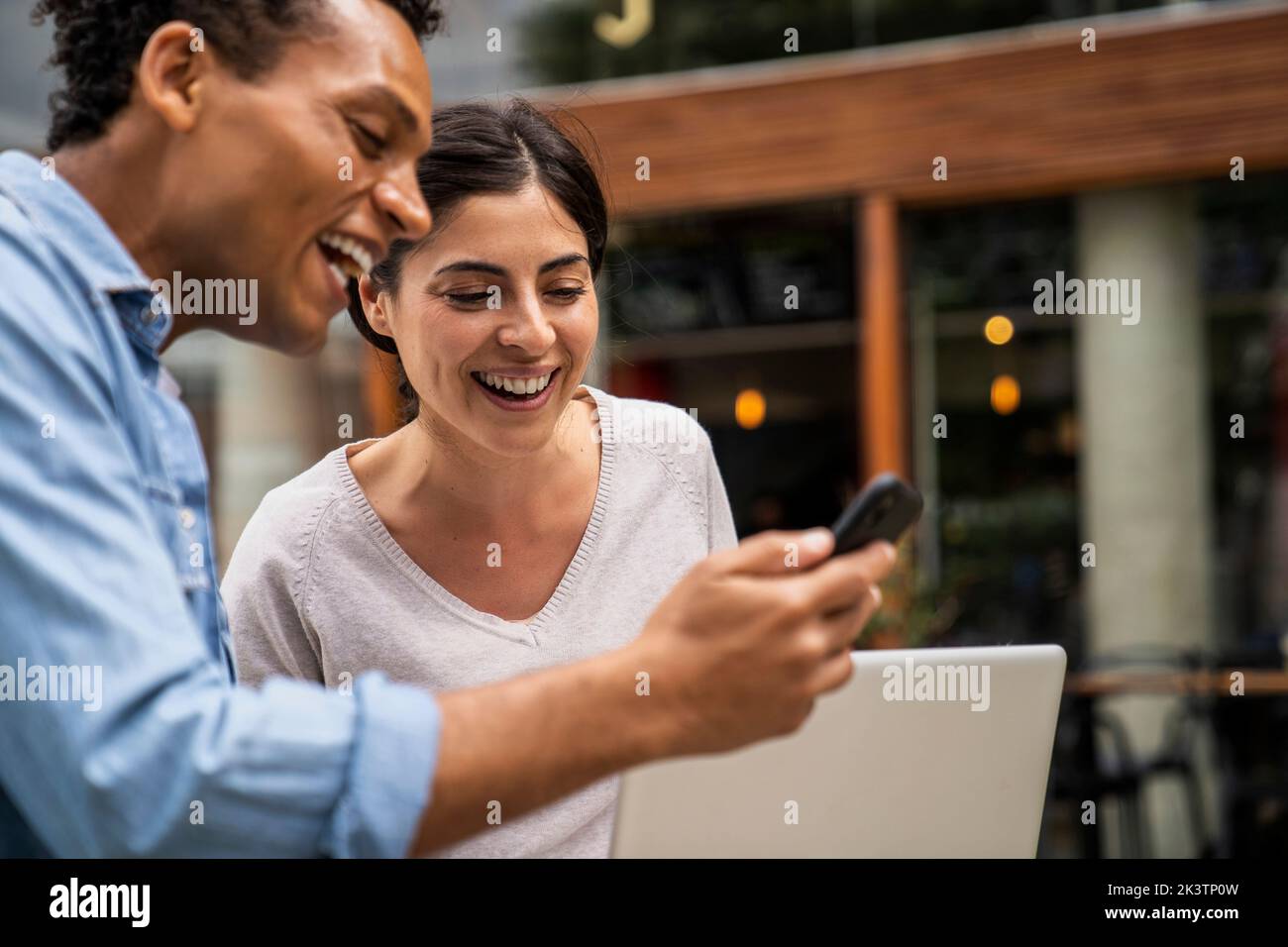 Seitenschuss afroamerikanischer Mann und lateinamerikanische Frau, die auf einen Smartphone-Bildschirm blicken und lachen Stockfoto