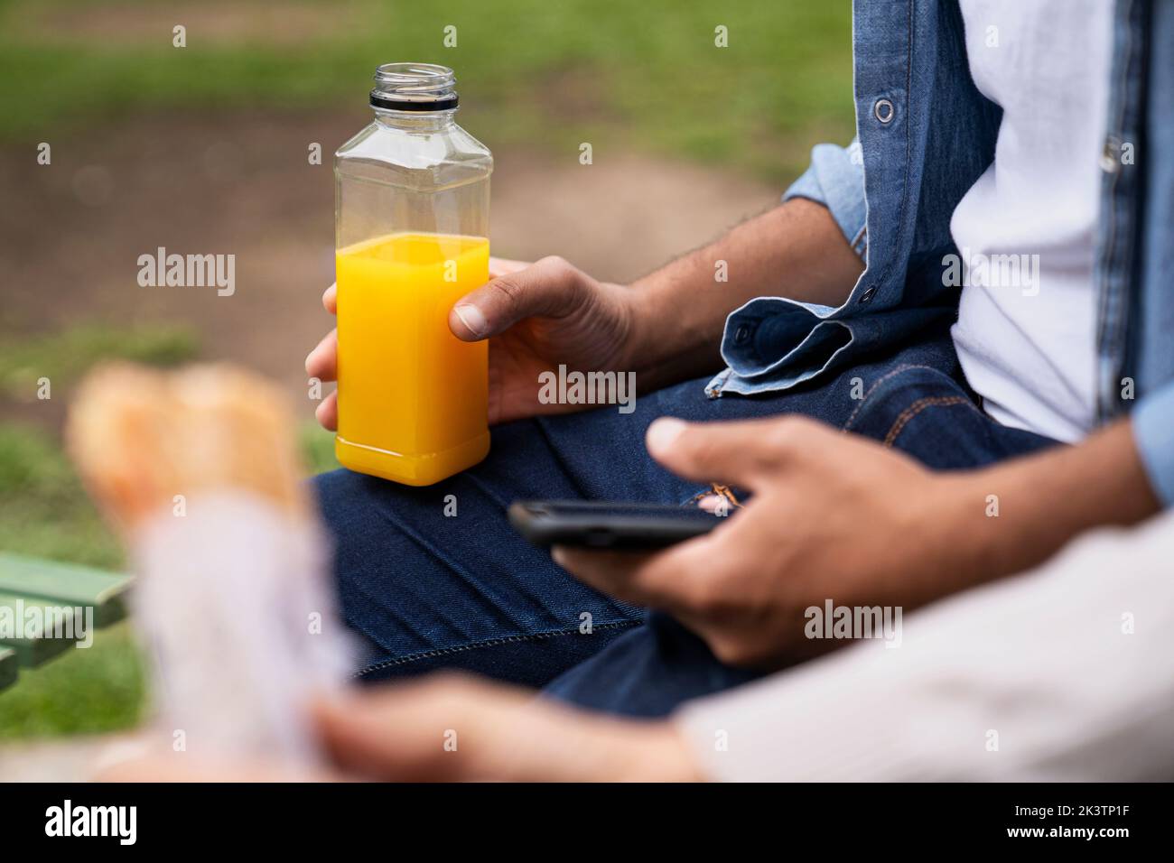 Mitten in der Hand eines Mannes, der eine Orangensaftflasche und eine unfokussiere Hand mit einem Sandwich hält Stockfoto
