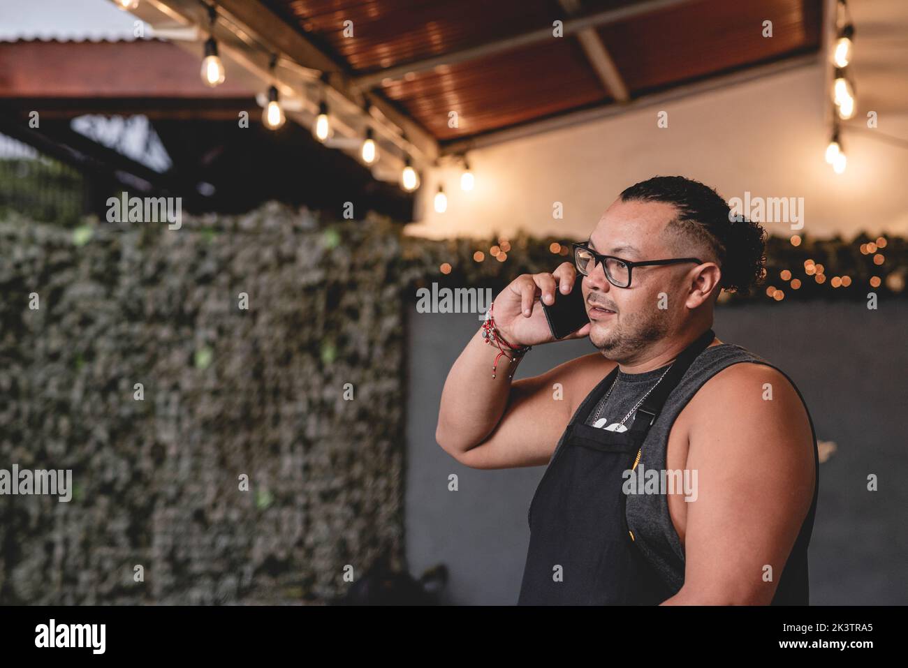 Ethnischer Mann, der auf dem Smartphone spricht, während er mit einer Flasche Bier auf dem Grillkocher mit offenem Deckel steht und Lebensmittel zubereitet Stockfoto