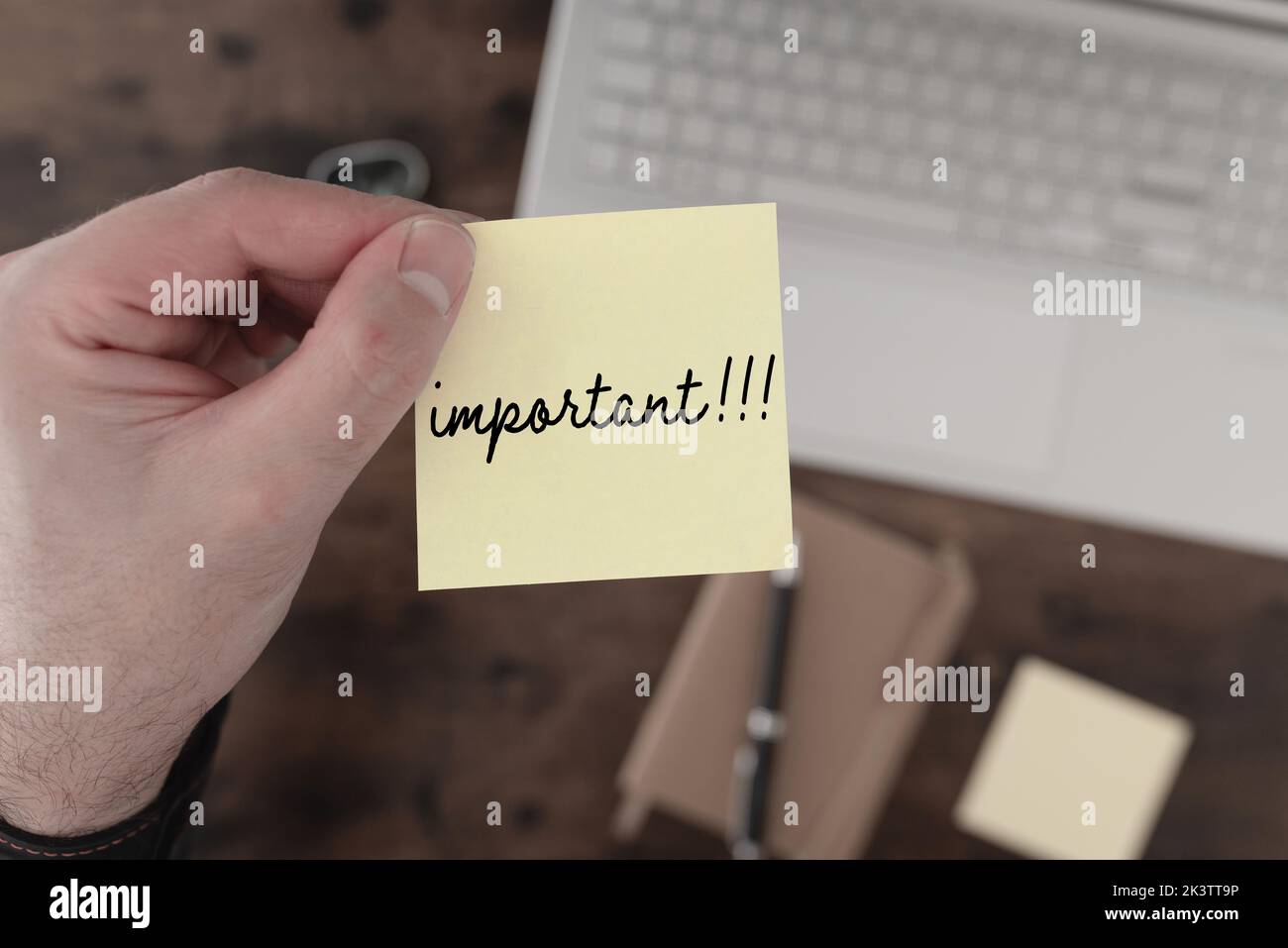 Draufsicht auf die Hand, die gelbe Haftnotizen mit dem Wort „WICHTIG“ über dem Schreibtisch mit Laptop und Notizblock hält Stockfoto