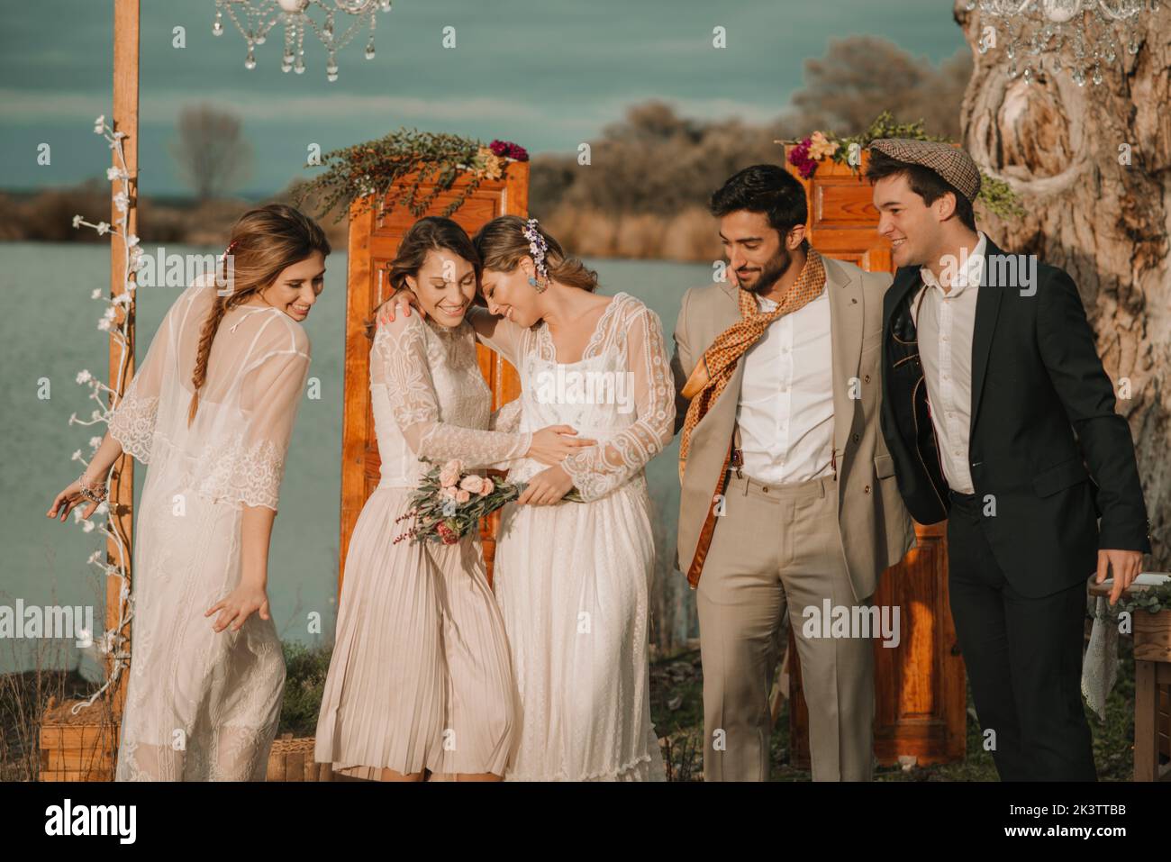 Junge lächelnde Jungs in Kostümen umarmt nahe attraktive fröhliche Damen in Kleidern mit Blumenstrauß in der Nähe von Dekorationen bei bewölktem Wetter Stockfoto