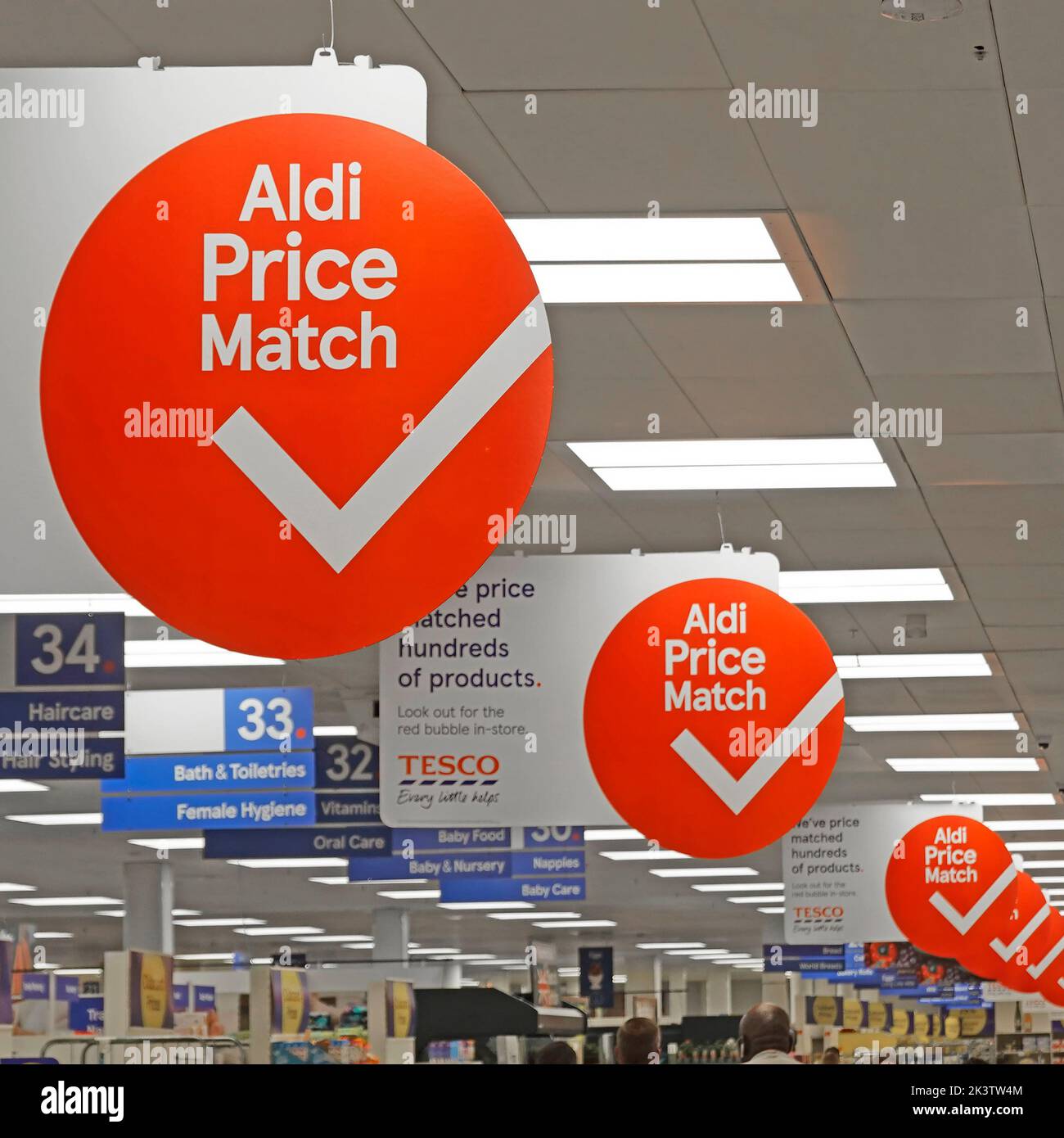 Repetitive runde Aldi Price Match Werbeschilder, die zwischen den Beleuchtungstafeln über dem Tesco Supermarkt-Inneneinkaufsgang Essex England UK hängen Stockfoto